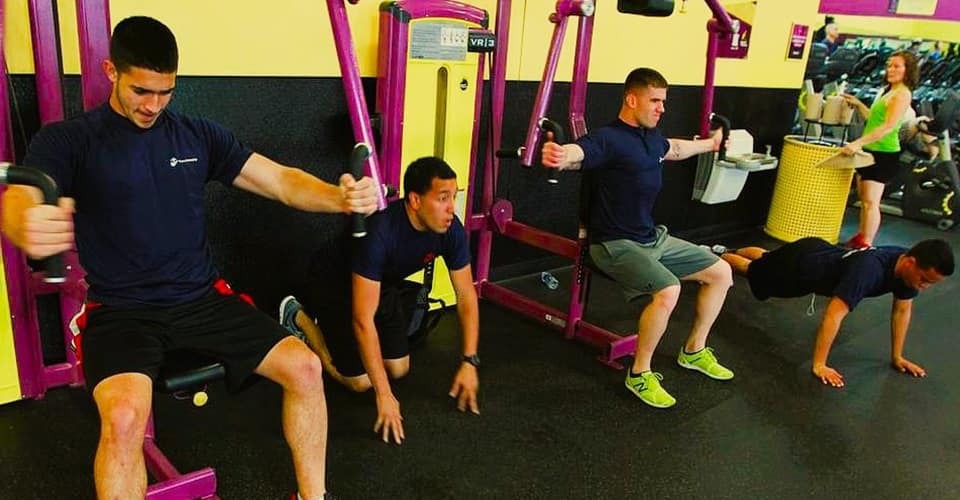 a row of young men working out at a fitness gym while a woman looks on