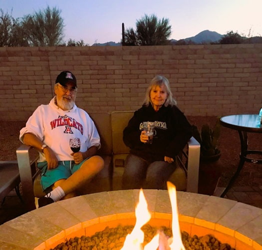 A couple holding drinks sitting next to a firepit in the early evening A couple holding drinks sitting next to a firepit in the early evening
