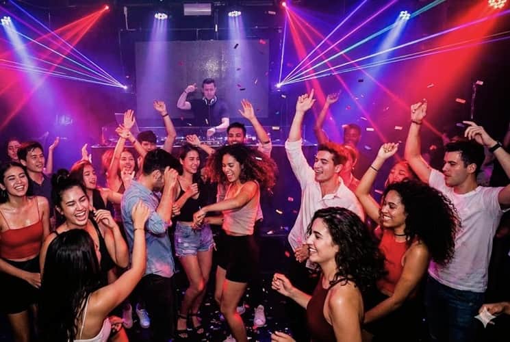 many young men and woman dancing to a dj in a club
